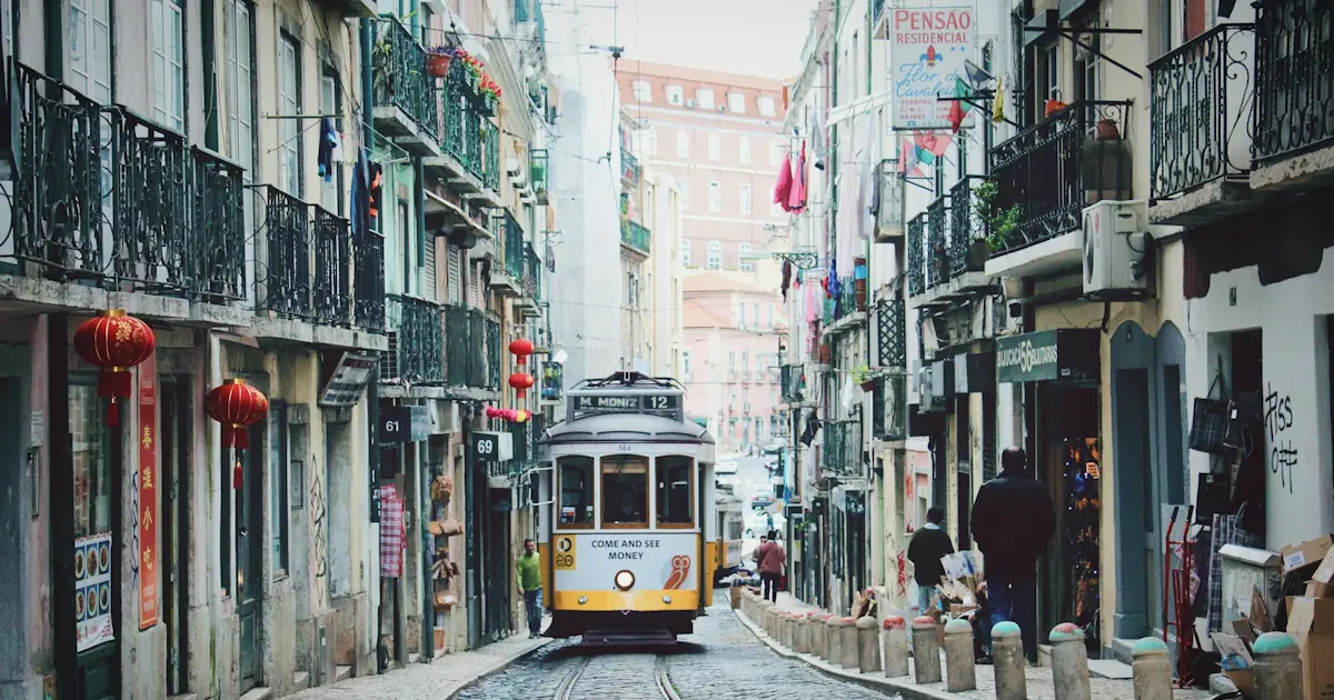 A yellow tram climbing a narrow cobblestone street in Lisbon on a sunny afternoon, with pastel-coloured buildings on both sides.