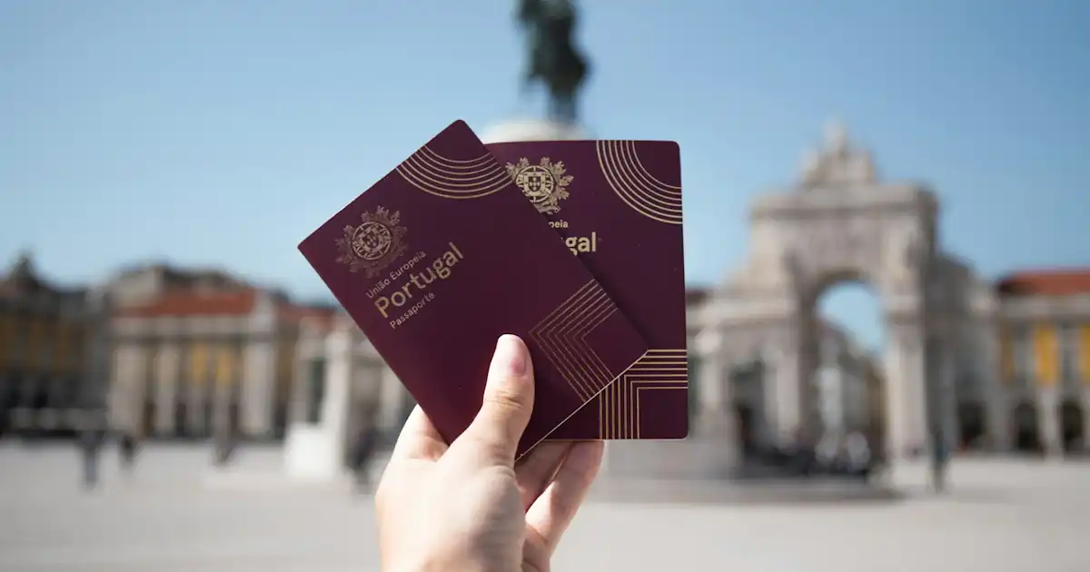 A hand is holding two Portuguese passports against the backdrop of Praça do Comércio in Lisbon, Portugal.